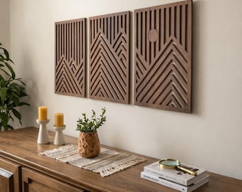 May include: Three rectangular dark brown wooden wall panels featuring vertical slats and a geometric mountain range design. The right panel includes a circular cutout representing a sun. The panels are mounted side-by-side on a light wall.