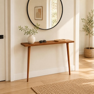 May include: A narrow wooden console table with four tapered legs. On the surface sits a white ceramic vase containing green leafy branches, a pair of black sunglasses, and a small oval wooden tray holding several small metal objects.