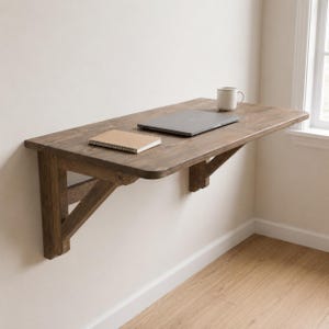 May include: A wall-mounted wooden desk with a dark brown finish, featuring two sturdy support brackets. A closed silver laptop, a small tan notebook, and a white ceramic mug are placed on the flat rectangular surface of the desk.