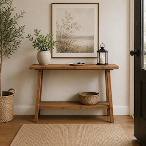 Può includere: Un tavolo console rettangolare in legno con ripiano inferiore, caratterizzato da una finitura marrone naturale. Il piano del tavolo ospita un vaso in ceramica bianca con fogliame verde, un piccolo piatto poco profondo e una lanterna nera.