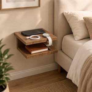 May include: A wall-mounted floating bedside table with a wood grain finish featuring two horizontal shelves. The top shelf holds a black smartphone, a white charging cable, and a wristwatch with a white band. A brown notebook rests on the bottom shelf.