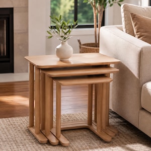 Può includere: Un set di tre tavolini a incastro in legno chiaro con superfici rettangolari lisce e gambe arrotondate. I tavolini sono impilati insieme, con un piccolo vaso in ceramica bianco sporco contenente rami verdi posizionato sul piano superiore.