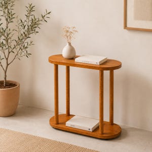 May include: A wooden console table with an oval top and base, supported by four cylindrical legs. The table features a smooth, warm-toned finish. A white vase with dried flowers sits on the top shelf, and a white book rests on the bottom shelf.