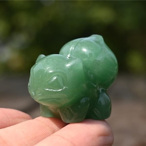 Può includere: Una piccola statuetta in pietra preziosa verde intagliata a forma di creatura con una struttura bulbosa sul dorso. La scultura presenta superfici lisce e levigate e un colore verde intenso traslucido, che cattura la forma di un personaggio.