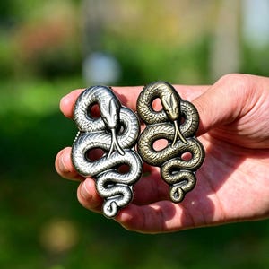 Peut inclure: Deux broches en métal en forme de serpent, l'une dans un ton argenté poli et l'autre dans un ton laiton antique. Chaque pièce présente un motif de serpent enroulé avec des écailles détaillées et une langue fourchue sortant de la tête.