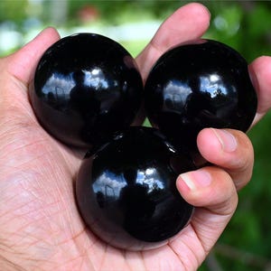 May include: Three smooth, polished, spherical black stones with a high-gloss finish. The spheres are dark and reflective, showing light highlights on their rounded surfaces. Each stone is perfectly round and solid black in colour, displaying a uniform appearance.
