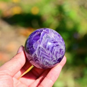 May include: A polished amethyst sphere, showcasing a deep purple hue with white and light purple crystalline patterns. The gemstone is held in a hand, with a blurred green and yellow background.