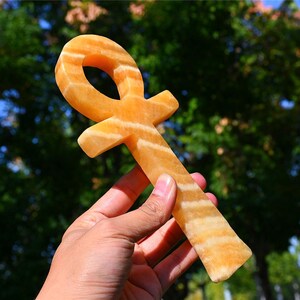 May include: A polished orange and white banded stone ankh symbol featuring a smooth, translucent texture with distinct horizontal layers of color throughout the cross-shaped object, held upright against a blurred outdoor green foliage background.