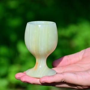May include: A small, pale green onyx wine glass with a rounded bowl and a short stem. The glass is held in a hand against a blurred green background. The glass has a striped pattern.