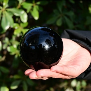 May include: A smooth, polished, spherical black stone orb with a highly reflective surface. The sphere is solid black in color and features a glossy finish that captures light reflections on its rounded exterior, showcasing a perfectly circular shape.