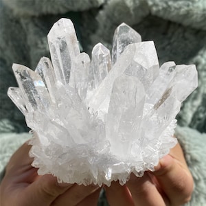 May include: A close-up of a cluster of clear quartz crystals. The crystals are tightly packed, with multiple pointed formations. The quartz has a natural, textured appearance. The background is soft and blurred, with the quartz held in a person's hands.