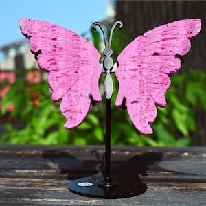 Puede incluir: Una escultura de mariposa rosa sobre un soporte de metal negro. Las alas de la mariposa tienen una superficie texturizada y son de color rosa brillante. La escultura se exhibe al aire libre, con un fondo verde borroso de follaje.