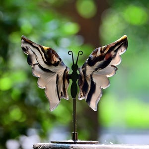 May include: A decorative butterfly sculpture with open wings, featuring a black and white striped pattern on the wings. The butterfly is mounted on a thin metal stand with a circular base. The background is a blurred green.