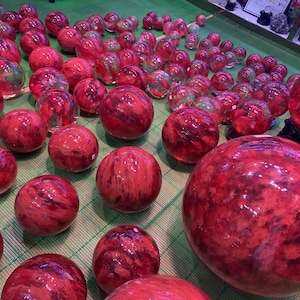 Puede incluir: Una gran colección de esferas de piedra o vidrio rojas y naranjas dispuestas sobre una superficie de rejilla verde. Las esferas varían en tamaño y presentan patrones de remolinos de color carmesí profundo, rojo brillante y naranja.