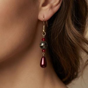 Puede incluir: Un pendiente largo y colgante con un gancho dorado. El pendiente presenta una cuenta en forma de lágrima de color rojo oscuro, una esfera gris oscuro y pequeñas cuentas rojas. El pendiente se lleva en la oreja de una persona.