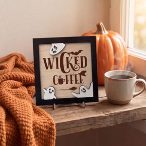 May include: A square wooden sign with a black frame featuring the text WICKED COFFEE in brown lettering. The design includes three white ghost cutouts, a small brown coffee cup icon, a flying bat silhouette, and a witch's broomstick graphic.