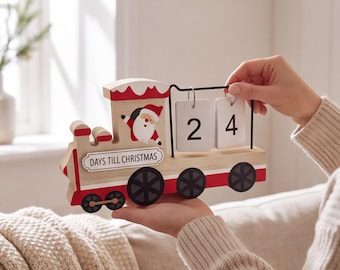 May include: A wooden train-shaped countdown calendar featuring a Santa Claus illustration and the text DAYS TILL CHRISTMAS. The train has red and white accents, black wheels, and two white hanging blocks displaying the number 24.