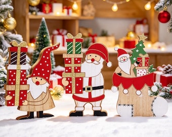 Op de afbeelding: Drie houten kersttafeldecoraties met een kabouter die gestapelde cadeaus vasthoudt, een kerstman met cadeaus en een kerstman in een kerstsok met een klein boompje, uitgevoerd in de kleuren rood, wit en groen.