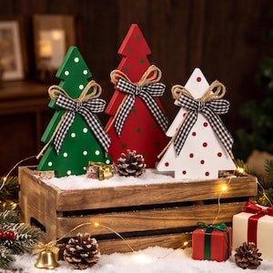 May include: Three wooden Christmas tree decorations in green, red, and white, each featuring polka dots and a black and white gingham ribbon bow with jute twine. The trees are arranged inside a rustic wooden crate filled with artificial snow.