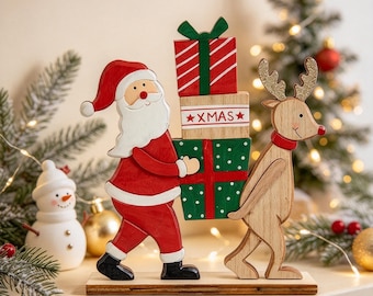 Puede incluir: Una decoración de mesa de madera que muestra a un Santa Claus pintado con traje rojo y un reno de madera natural cargando una pila de tres cajas de regalo. La caja central es blanca con el texto rojo XMAS y dos estrellas rojas a los lados.