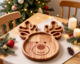 May include: A light brown wooden serving tray shaped like a reindeer head with prominent antlers. The tray features a smooth finish, a circular face area, two small eyes, and a darker wood inlay for the nose and a curved mouth line.