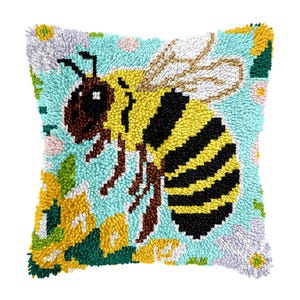 May include: Square decorative pillow with a large bee design. The bee is yellow and black, set against a light blue background with floral accents in yellow, green, and purple. The pillow has a textured, handmade appearance.