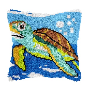 May include: Square, handmade pillow featuring a colorful sea turtle design. The turtle has a brown shell, green and yellow flippers, and a turquoise body. The background is blue with white accents, creating a sea and sky scene. The pillow is made of a textured material.