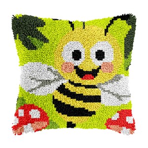 Könnte beinhalten: Quadratisches, handgefertigtes Kissen mit einem Cartoon-Bienen-Design. Die Biene hat einen gelben Körper mit schwarzen Streifen, weiße Flügel und ein lächelndes Gesicht. Der Hintergrund ist grün mit roten und weißen Pilzakzenten. Das Kissen ist aus einem strukturierten, geschlungenen Stoff.