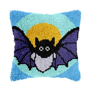 May include: A square punch needle throw pillow featuring a black bat with white eyes and a white chest. The bat is centered against a bright yellow circular moon, which is set against a light blue background with dark blue bat wings.