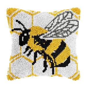 Könnte beinhalten: Quadratisches Zierkissen mit einem detaillierten gestickten Bienenmuster. Die Biene ist gelb und schwarz, vor einem weißen Hintergrund mit einem gelben Wabenmuster. Das Kissen hat ein strukturiertes, weiches Aussehen.