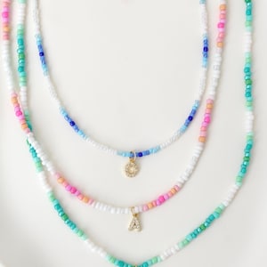 Könnte beinhalten: Drei Perlenketten mit bunten Perlen in Weiß-, Blau-, Rosa- und Grüntönen. Jede Halskette hat einen goldfarbenen Initialenanhänger. Die Halsketten werden auf einer weißen Oberfläche präsentiert.