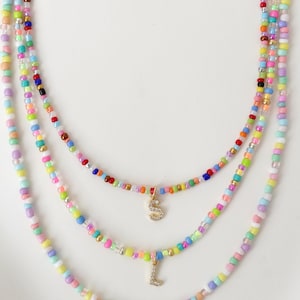 May include: Three layered beaded necklaces in pastel shades. Each necklace has small, colourful beads and a gold-toned charm with a letter. The letters are S, L, and I. The necklaces are displayed on a white surface.
