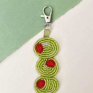 May include: A beaded keyring featuring three stacked green olives with red pimentos, crafted from small glass beads in shades of lime green, white, and red. The design is attached to a silver-toned metal lobster clasp by a short green fabric loop.