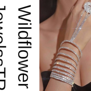 Puede incluir: Una brillante pulsera de cadena de mano plateada con múltiples filas de brillantes pedrería. La pulsera se conecta a un anillo con un gran diseño circular de pedrería. El texto "JewelsSTR Wildflower" es visible en la imagen.