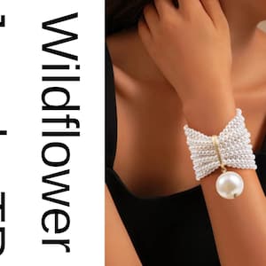 Könnte beinhalten: Ein weißes Perlenarmband mit Schleifendesign und einer großen, hängenden Perle. Das Armband besteht aus kleinen, runden Perlen und hat ein goldfarbenes Band. Der Text "Wildflower Jewels TR" ist sichtbar.