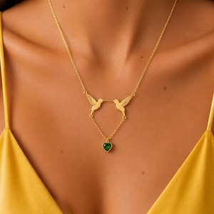 May include: A gold-toned necklace featuring two hummingbird pendants facing each other, connected by a delicate chain that suspends a small, faceted, heart-shaped green gemstone charm at the center point of the design.