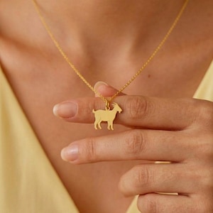 May include: A gold-colored necklace featuring a goat pendant. The goat silhouette is detailed and hangs from a delicate chain. The necklace is displayed against a soft yellow background.