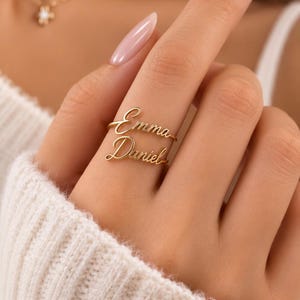 Op de afbeelding: Een goudkleurige ring met twee gestapelde naamplaatjes in cursief schrift. Het bovenste naamplaatje toont de naam Emma en het onderste naamplaatje toont de naam Daniel. De ring is gemaakt van gepolijst metaal met een warme metallic afwerking.