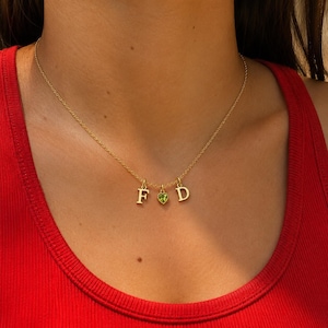 May include: A gold-toned chain necklace featuring three small dangling charms: a capital letter F, a central heart-shaped green gemstone, and a capital letter D. The necklace rests against the neck, showcasing a delicate link design and polished metal.