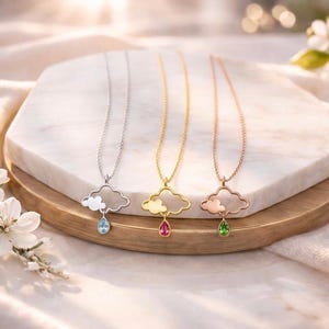 Puede incluir: Tres collares con colgantes en forma de nube con gemas en forma de lágrima. Los collares son plateados, dorados y oro rosa, cada uno con una gema de diferente color: azul, rosa y verde. Los collares se exhiben sobre una superficie de mármol y madera.