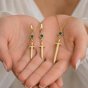 Könnte beinhalten: Ein goldfarbenes Schmuckset bestehend aus zwei Creolen und einer Anhängerkette. Jedes Teil zeigt einen kleinen runden grünen Edelstein, der über einem goldenen Schwertanhänger sitzt. Die Kette besteht aus einer feinen goldenen Gliederkette.