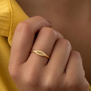 以下が含まれることがあります： 滑らかな楕円形のフェイスプレートを備えたゴールドトーンのメタルリング。フェイスプレートの中央には、クリアでダークなサンセリフフォントで「Sophia」という名前が刻印されています。リングのバンド部分は細く、指の周りで一定の幅を保っています。