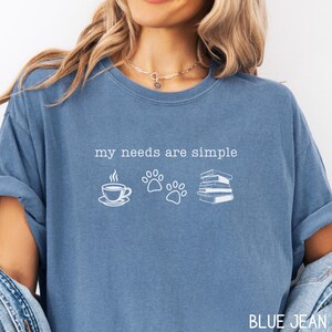 May include: Blue jean colored t-shirt with the text "my needs are simple" above a coffee cup, paw prints, and a stack of books. The shirt is a solid color with a relaxed fit.