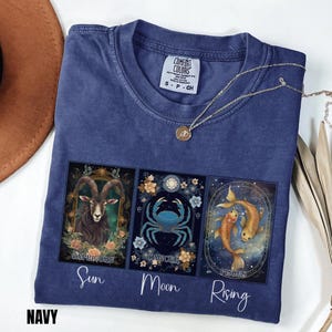 May include: A navy blue t-shirt featuring three rectangular panels with zodiac illustrations of a goat for Capricorn, a crab for Cancer, and two fish for Pisces. Below the panels, the words Sun, Moon, and Rising are printed in white script.