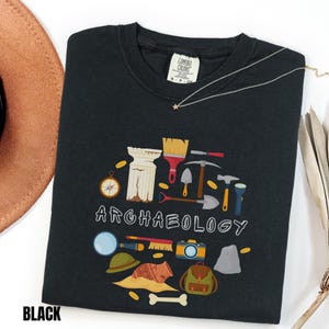 Peut inclure: Un t-shirt noir avec une impression graphique représentant divers outils et artefacts archéologiques, dont une colonne, un pinceau, une pioche, un marteau, une loupe, un appareil photo et un sac à dos, avec le mot ARCHAEOLOGY en lettres blanches.
