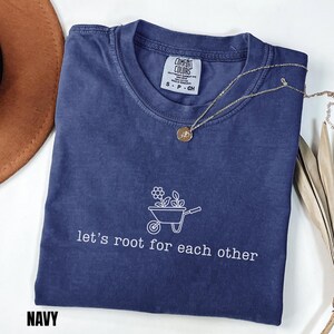 May include: Navy blue t-shirt featuring the text "let's root for each other" and a wheelbarrow graphic with flowers. The shirt has a crew neck and is made of a soft material. A gold necklace with a pendant is on the shirt.