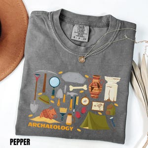 Peut inclure: Un t-shirt gris anthracite avec une impression graphique d'outils et d'artefacts archéologiques, incluant une pioche, une pelle, une loupe, de la poterie, un os, une boussole, une carte et une tente, avec le texte ARCHAEOLOGY imprimé en jaune.