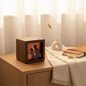 Peut inclure: Cadre photo en bois en forme de cube, affichant une image rétroéclairée d'un couple. Le cadre est en bois foncé et posé sur une surface en bois. Un petit vase blanc et un carnet en cuir sont également visibles.