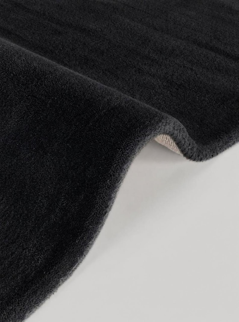 May include: A thick, charcoal gray rug with a soft, plush, high-pile texture. The rug is folded over to reveal a light-colored, woven backing material underneath the dense, dark-colored fibers that cover the entire top surface.