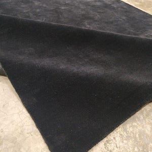 May include: A rectangular area rug with a solid black, dense, and short-pile surface. The rug is shown partially folded over, revealing the dark texture of the material and its straight, finished edges against a light-colored floor.